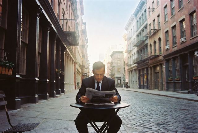 7:25 AM • Financial District — Stone Street Quiet: A lone suited man reads the newspaper at a small outdoor table on historic cobblestone before the Wall Street rush.